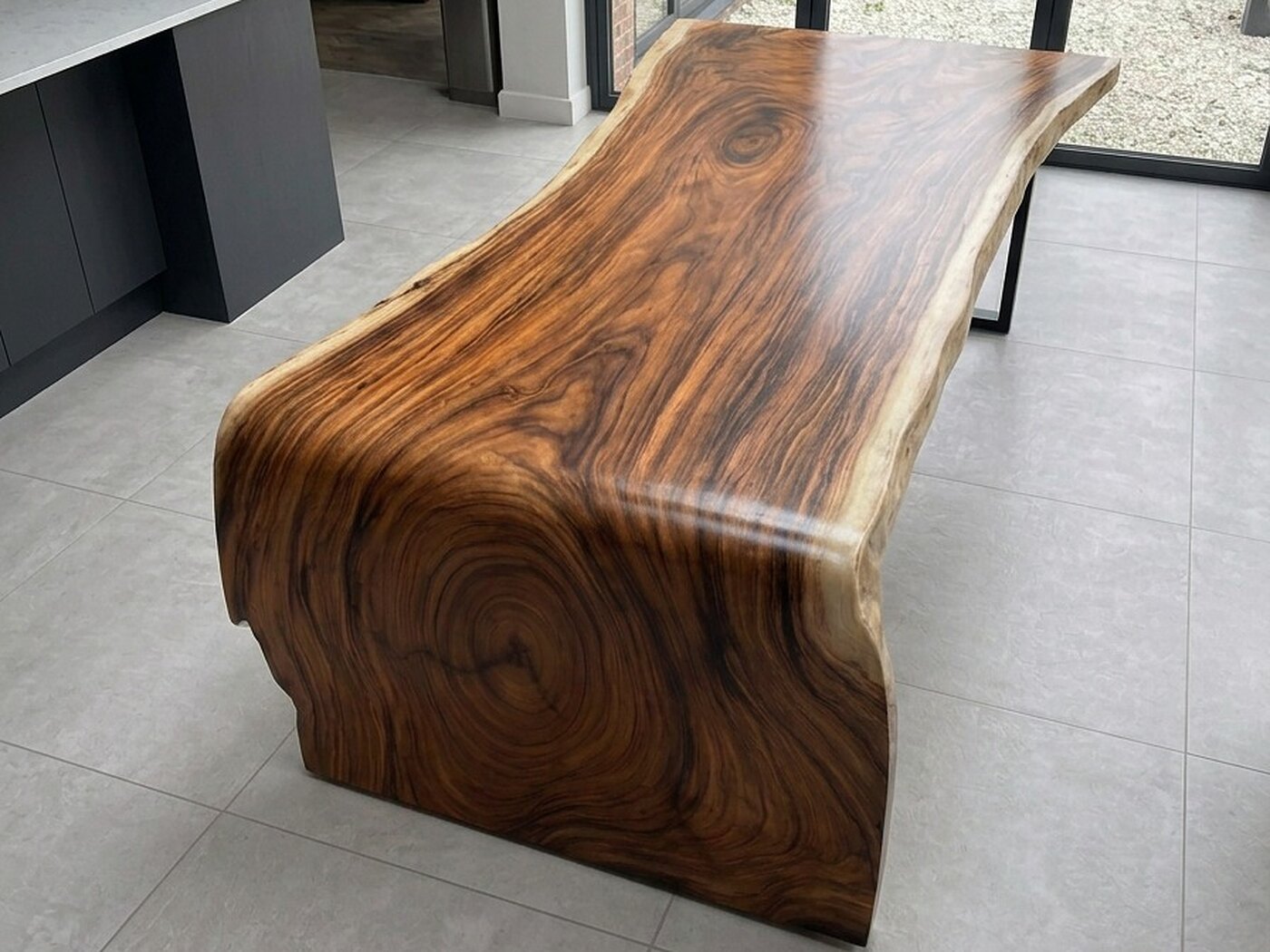 The table closer in, the rings of the slab opening across the face of its fall, charcoal kitchen units to the left and a slim black steel leg at the far end
