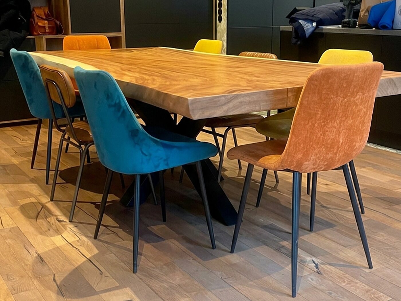 A bright honey Suar table in a kitchen, surrounded by velvet chairs in teal, mustard, orange and tan, with dark tall units and oak-lined cubbies behind