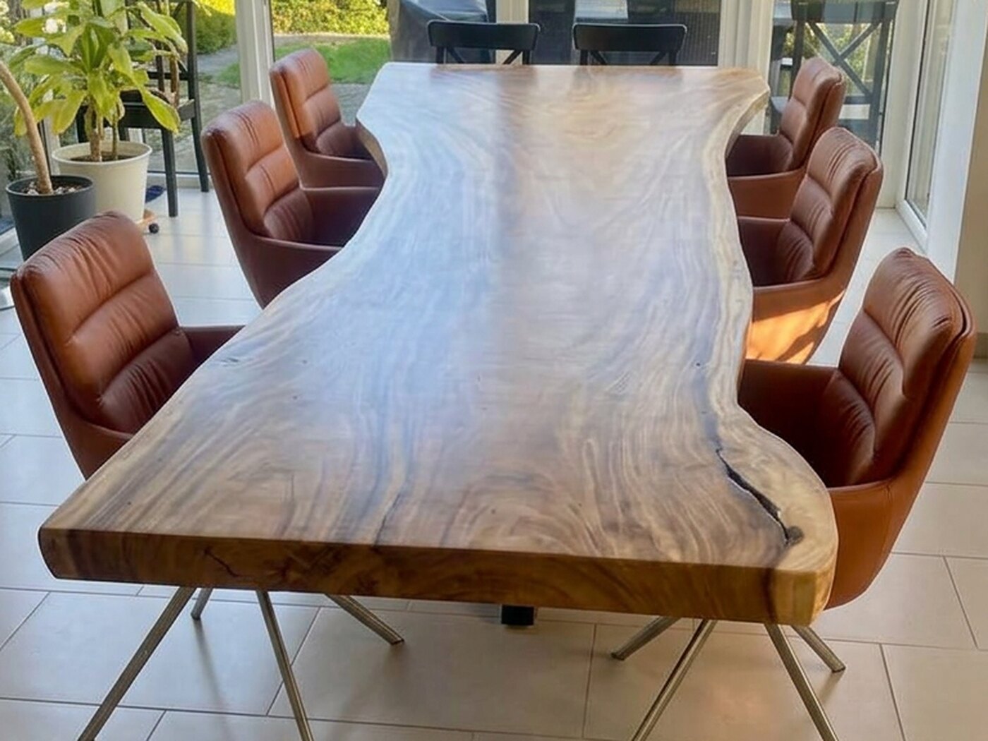 A long live-edge Suar table with tan leather chairs down both sides, standing in a glazed garden room with a clipped hedge and a terrace beyond