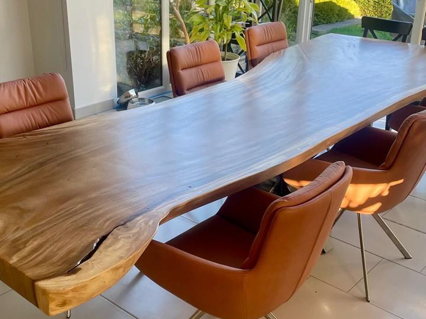 A live-edge Suar table seen down its length in a glazed garden room, a roller blind above and planting outside