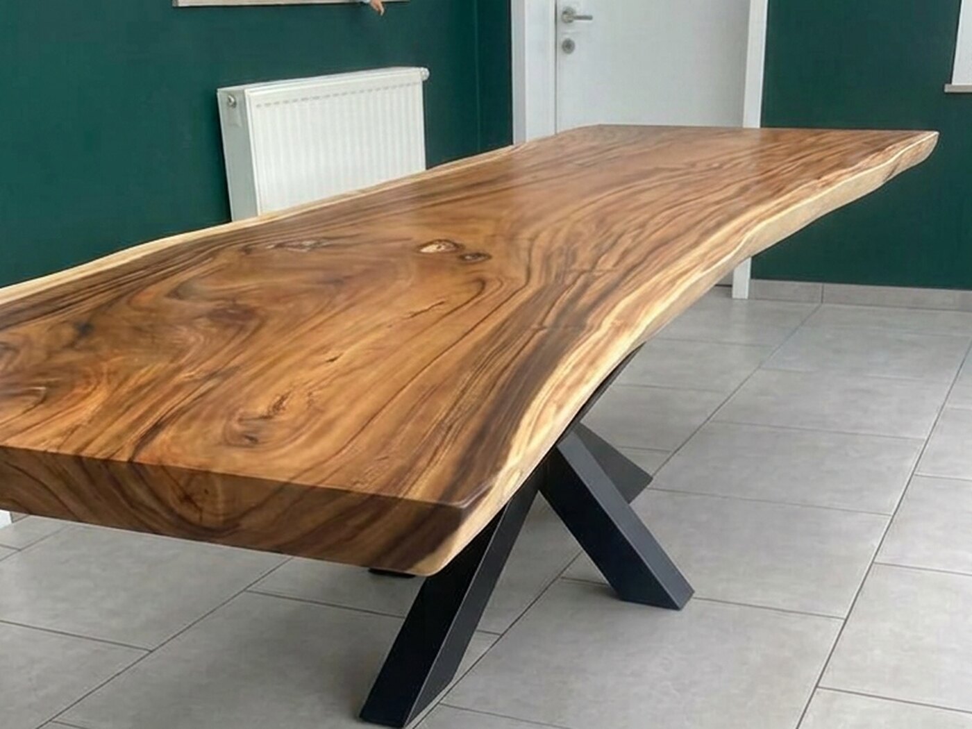 A live-edge Suar table on a black crossed frame, standing on grey floor tiles against a wall painted deep green, the room still unfinished