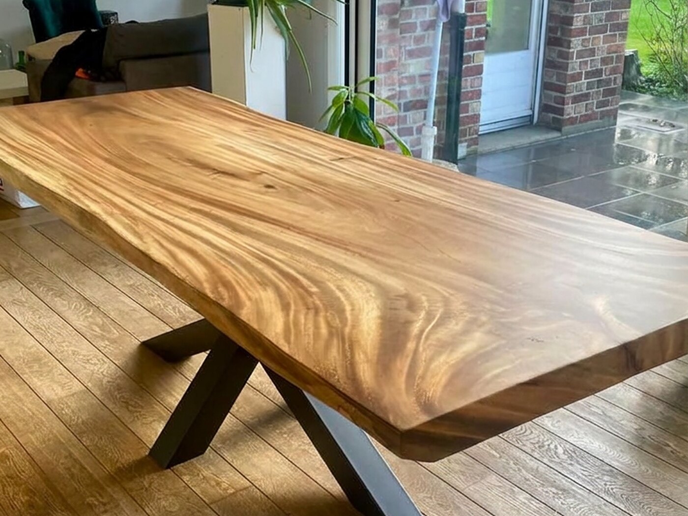 A honey-coloured live-edge Suar table on a black spider frame, standing on wide oak boards with sliding glass onto a brick courtyard behind it