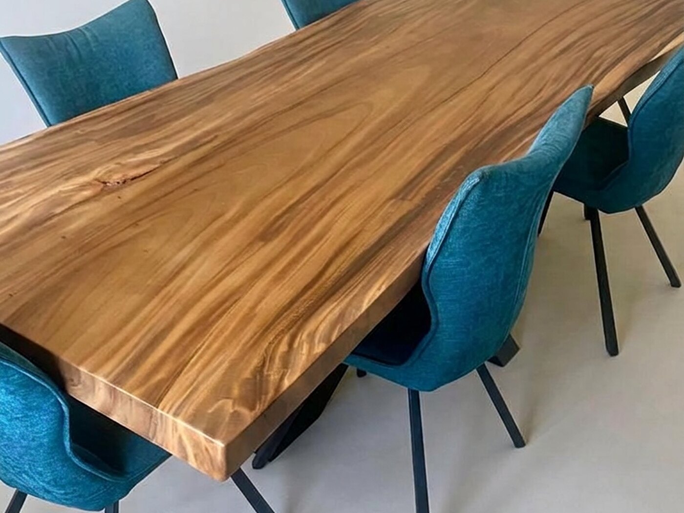 A golden live-edge Suar table with teal blue chairs down both sides, standing alone in a bare cream-walled room