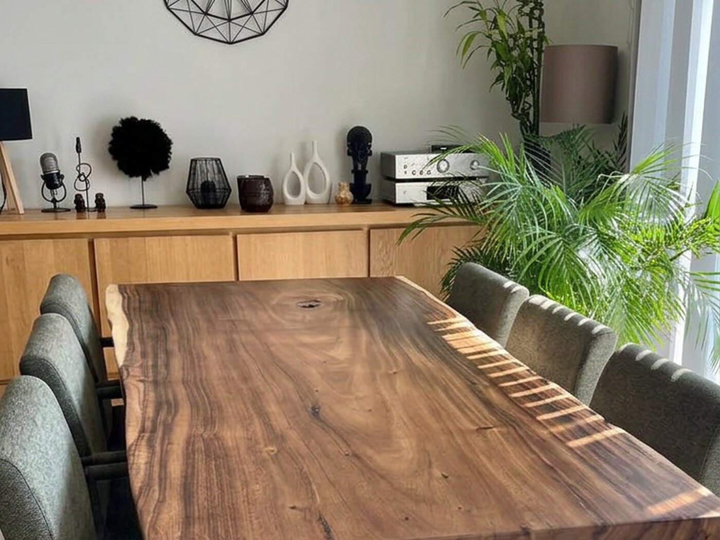 A deep brown live-edge Suar table under three black wire-cage pendants, with grey-green chairs down both sides and an oak sideboard behind