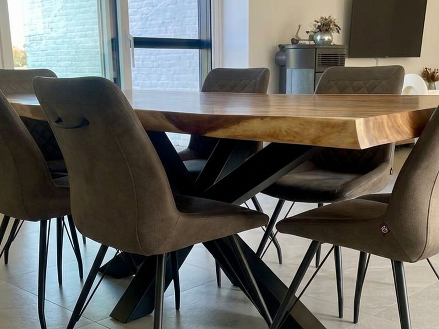 A warm red-brown live-edge Suar table on a black spider frame, with brown upholstered chairs around it, tall windows to the left and a freestanding stove beyond