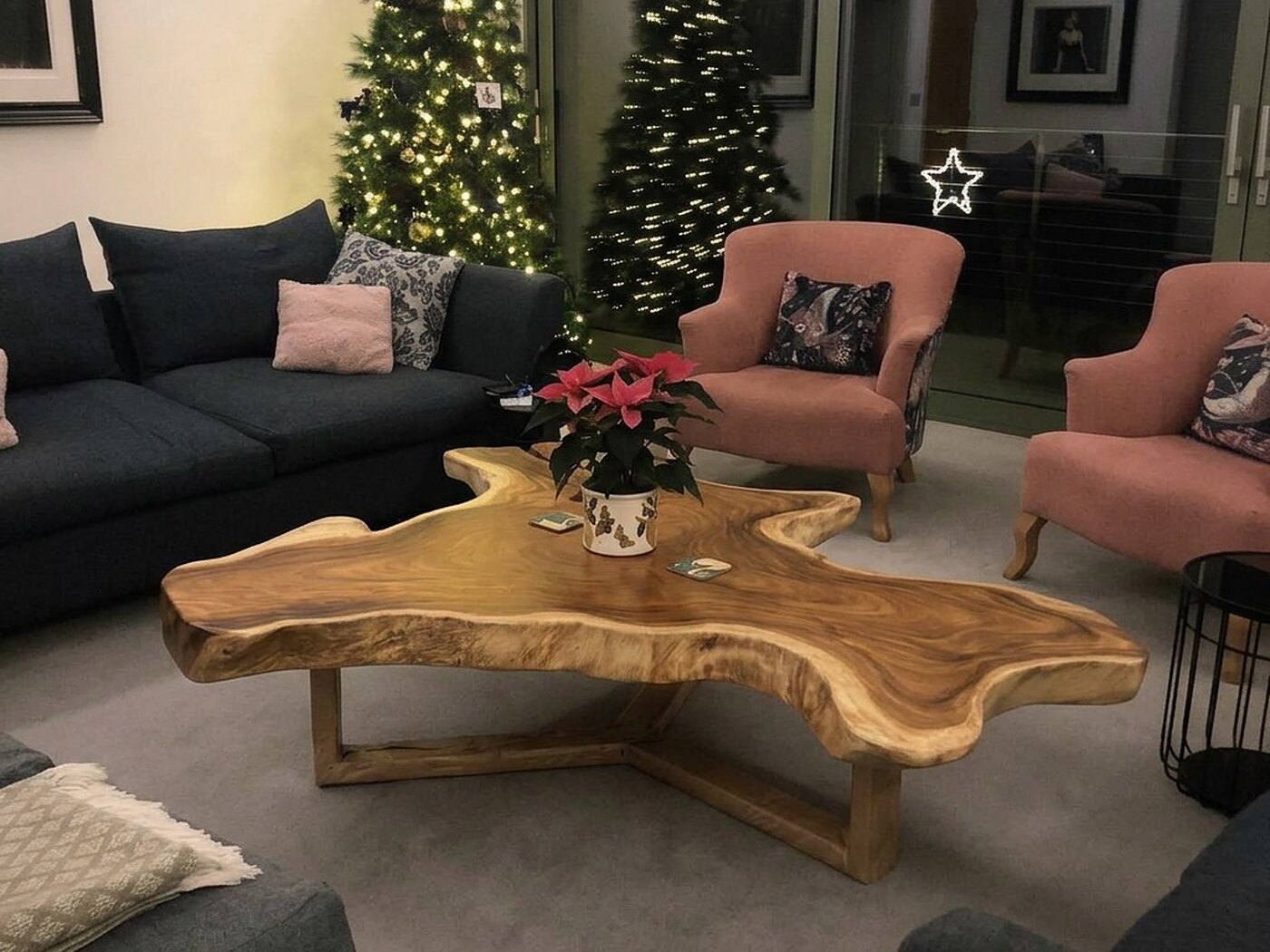 The table across the room at night, charcoal sofas to the left, two pink armchairs behind it and a lit Christmas tree against the glass