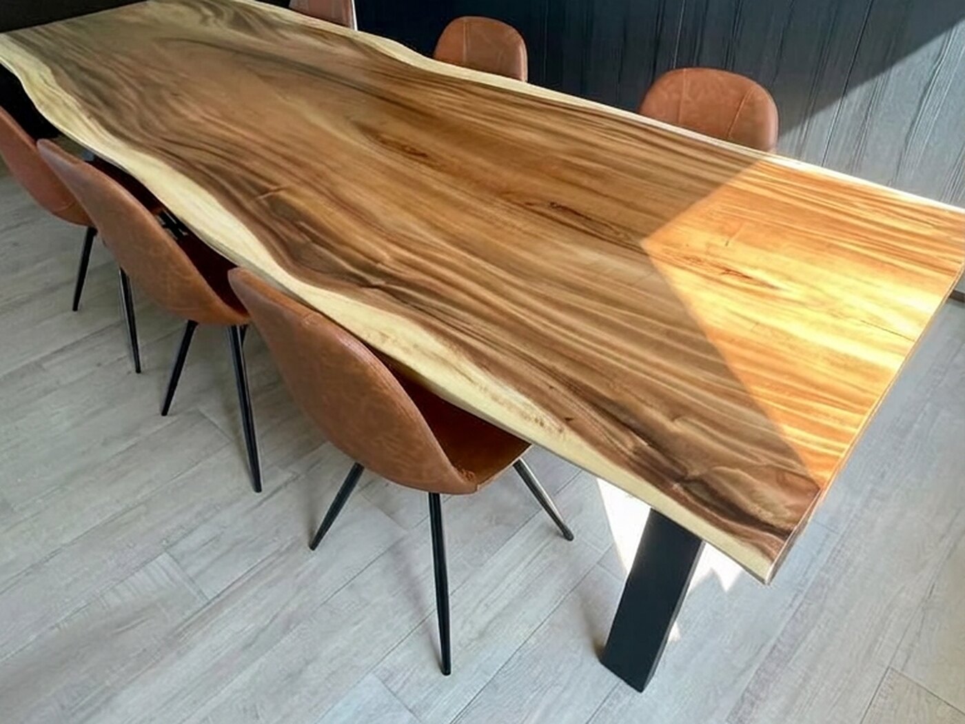A live-edge Suar table with tan leather chairs, standing against a wall of near-black vertical panelling with an oak-lined picture recess